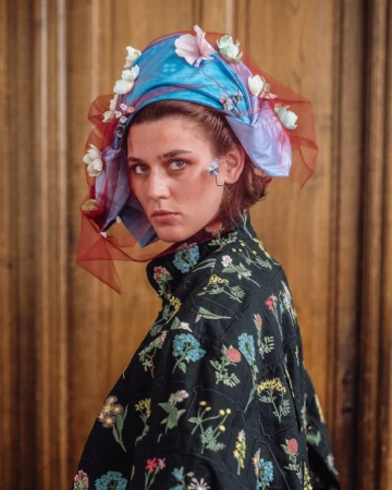Model wearing a colourful floral headwrap and embroidered black jacket looking over shoulder against wood paneling