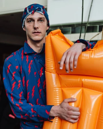 Model leaning on a bright orange inflatable object wearing a blue lightning-bolt patterned shirt and matching cap