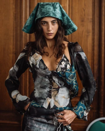 Model wearing a teal floral hat and patterned blouse in a backstage setting captured by Giacomo Cosua.