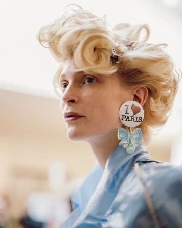 Model with voluminous blonde hair and large I Love Paris statement earrings wearing a blue garment backstage.