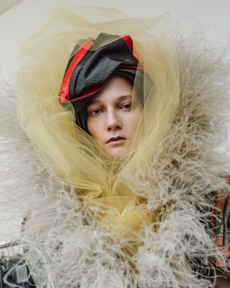 Model wearing feathered collar and yellow tulle headpiece backstage at Vivienne Westwood S/S 27