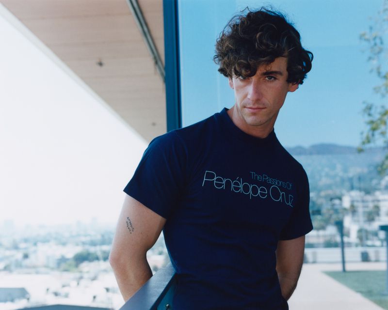 Young man with curly dark hair wearing a dark blue t-shirt leaning against a balcony railing outdoors