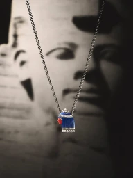 Close-up jewellery product shot of a silver chain necklace with a pharaoh pendant featuring blue and red enamel against a dark background