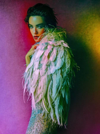 Model wearing a feathered cape and embellished dress against a colorful gradient background