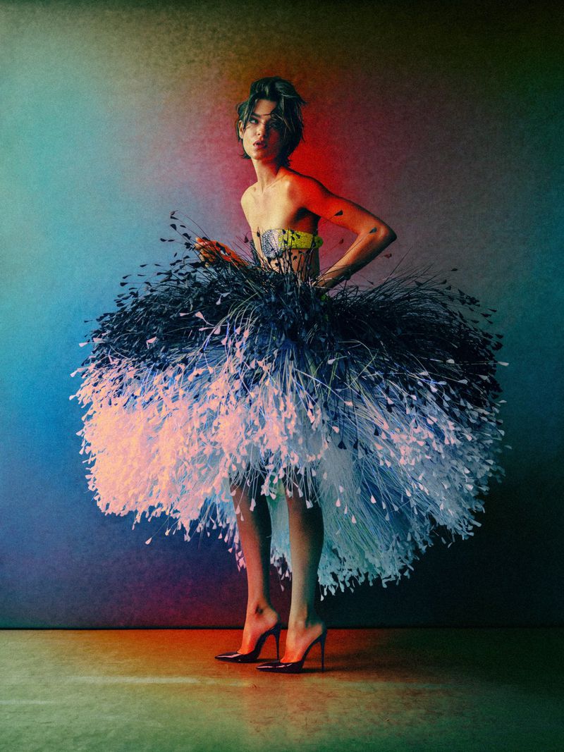 Model in voluminous black and white feather dress wearing high heels against a multi-coloured gradient background.