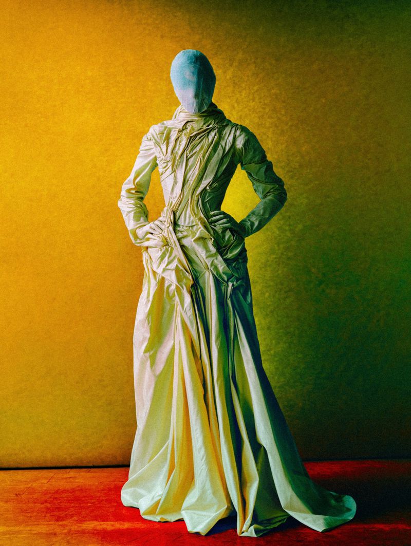 Model in sculpted light yellow gown with face covered by fabric standing against a gold and green textured background.