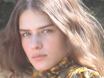 Close-up portrait of a model with long brown hair wearing a plaid top against an outdoor background