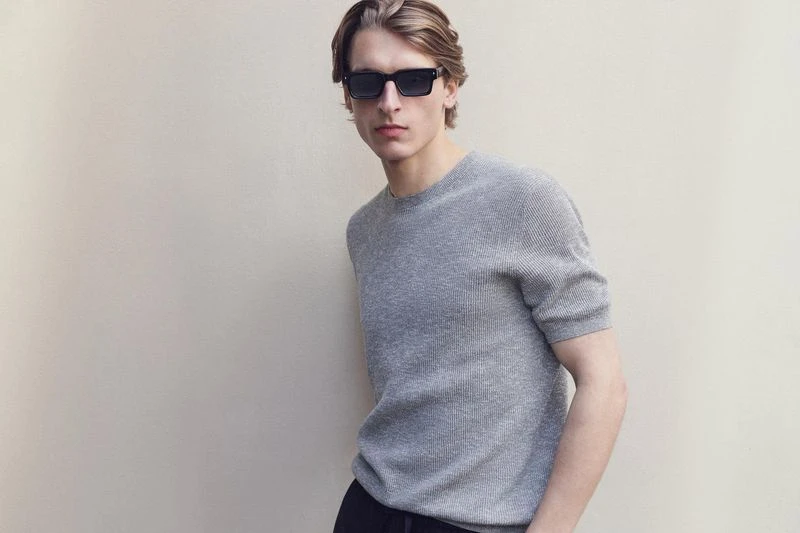 A young adult male poses in a gray ribbed, short-sleeved shirt and sunglasses against a neutral backdrop, creating a fashionable, modern look.