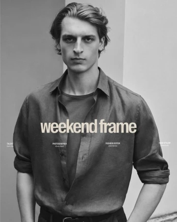 Portrait of a young adult male fashion model in a casual linen shirt and dark pants posing against a grey and off-white wall indoors.
