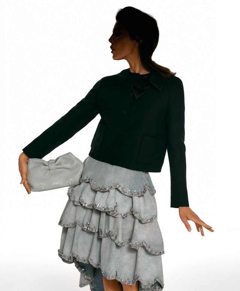 Monica Barbaro in dark jacket and layered ruffle skirt holding a light grey clutch against white seamless background