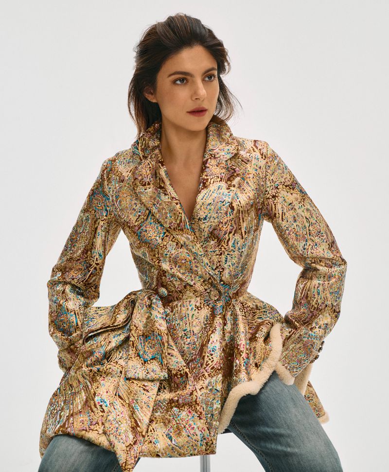 Monica Barbaro wearing a patterned patterned belted jacket and blue jeans against a clean white backdrop
