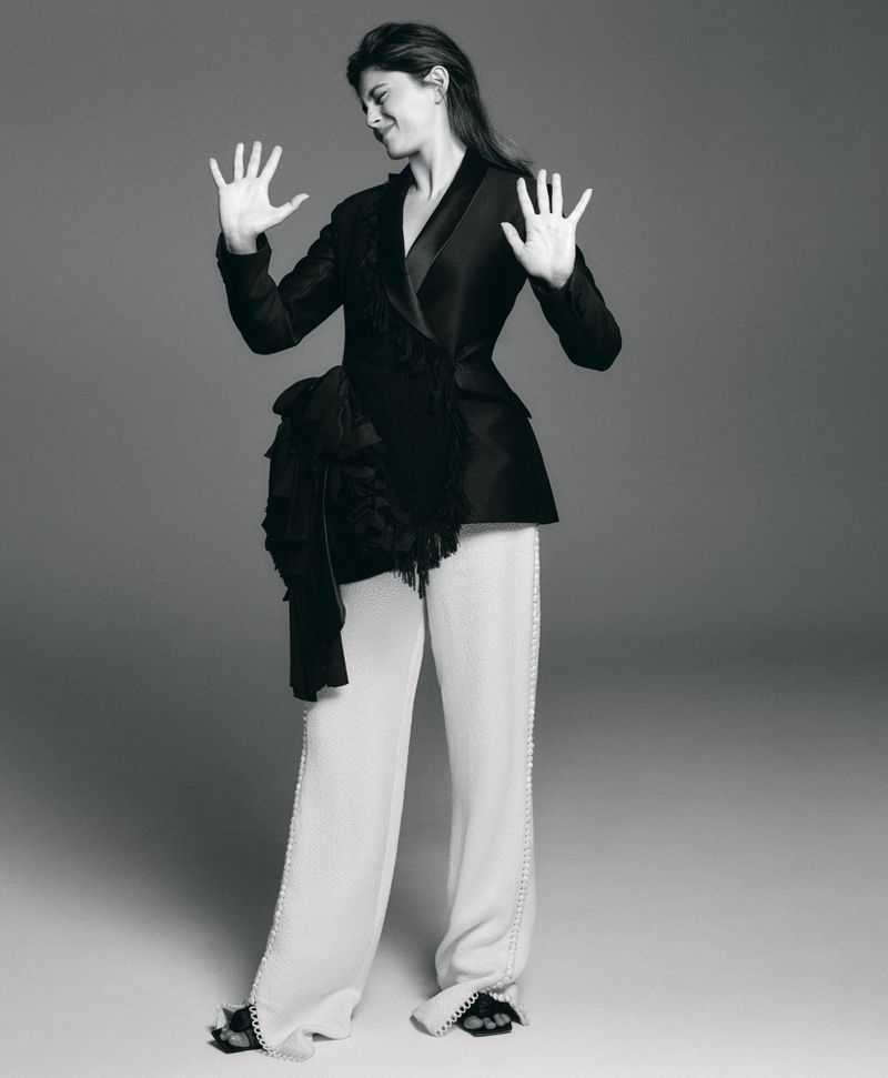 Monica Barbaro in a black jacket and white trousers with hands raised against a grey background