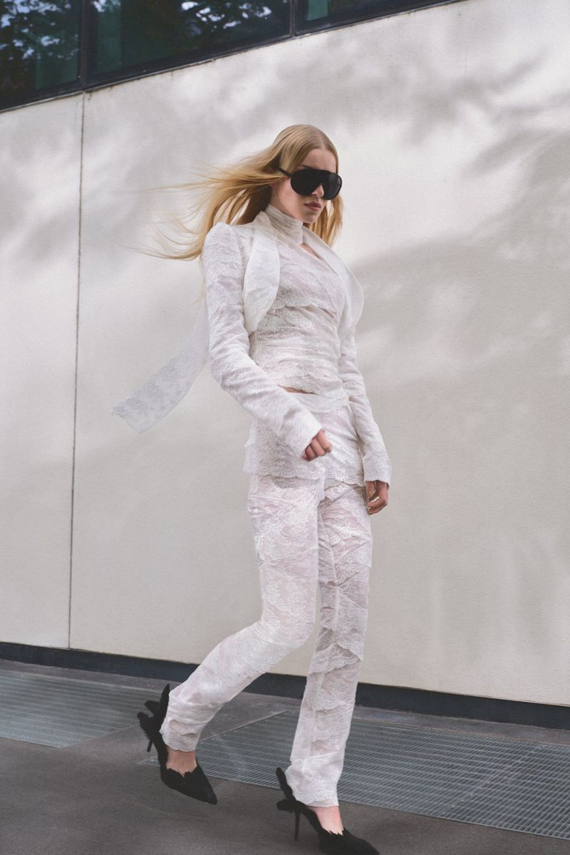 Model in white lace suit and dark sunglasses walking outdoors against a building exterior wall