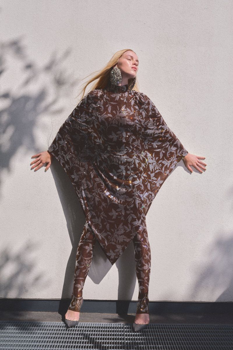 Model in a brown patterned tunic and matching leggings posing against a white exterior wall with leaf shadows