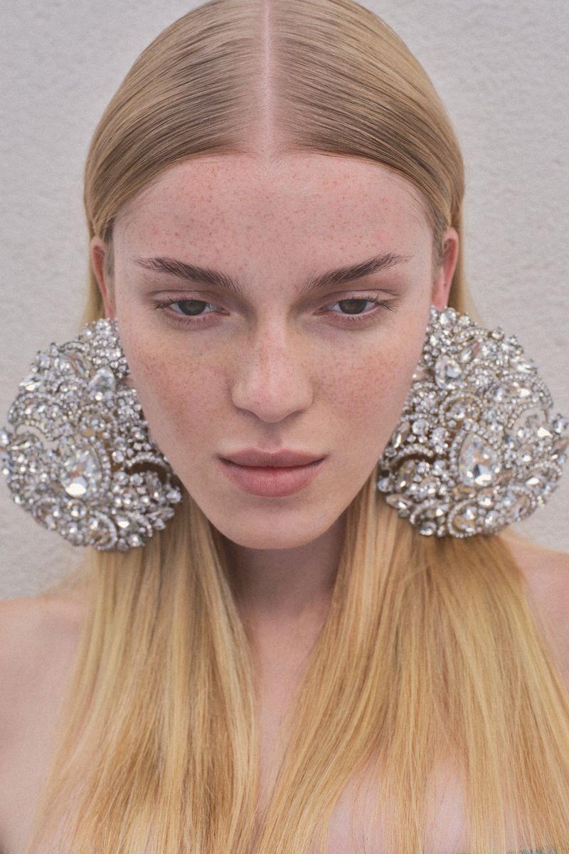 Close-up portrait of a woman with long blonde hair and freckles wearing large crystal embellished statement earrings.