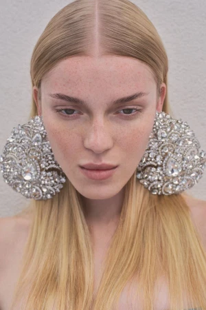 Close-up portrait of a woman with long blonde hair and freckles wearing large crystal embellished statement earrings.