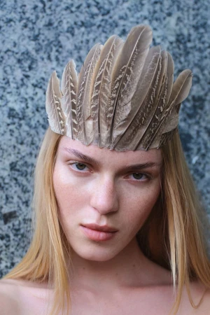 Close-up portrait of a model wearing a structured feathered crown against a grey stone wall background.