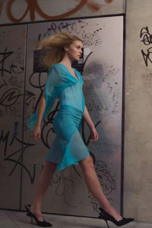 Model walking past a graffiti-covered wall wearing a sheer turquoise dress and black heels.