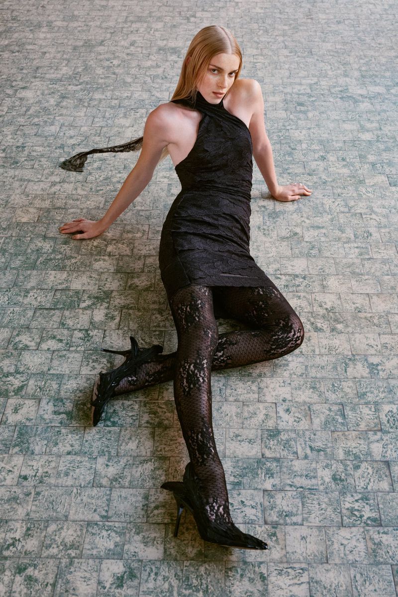 Model seated on a patterned tiled floor wearing a black halterneck dress and patterned black tights.