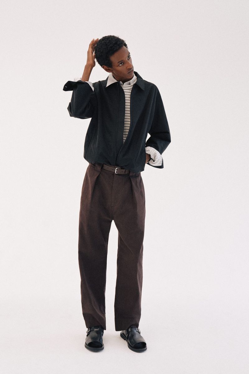 Model wearing dark jacket, striped shirt, and brown trousers posing against a plain white background
