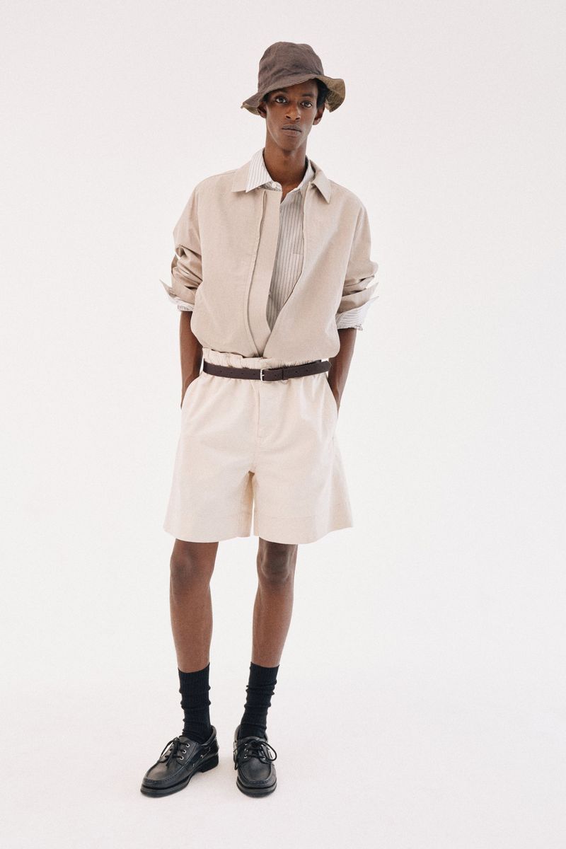 Model wearing a bucket hat, beige collared shirt, matching shorts, and dark shoes against a plain white background.