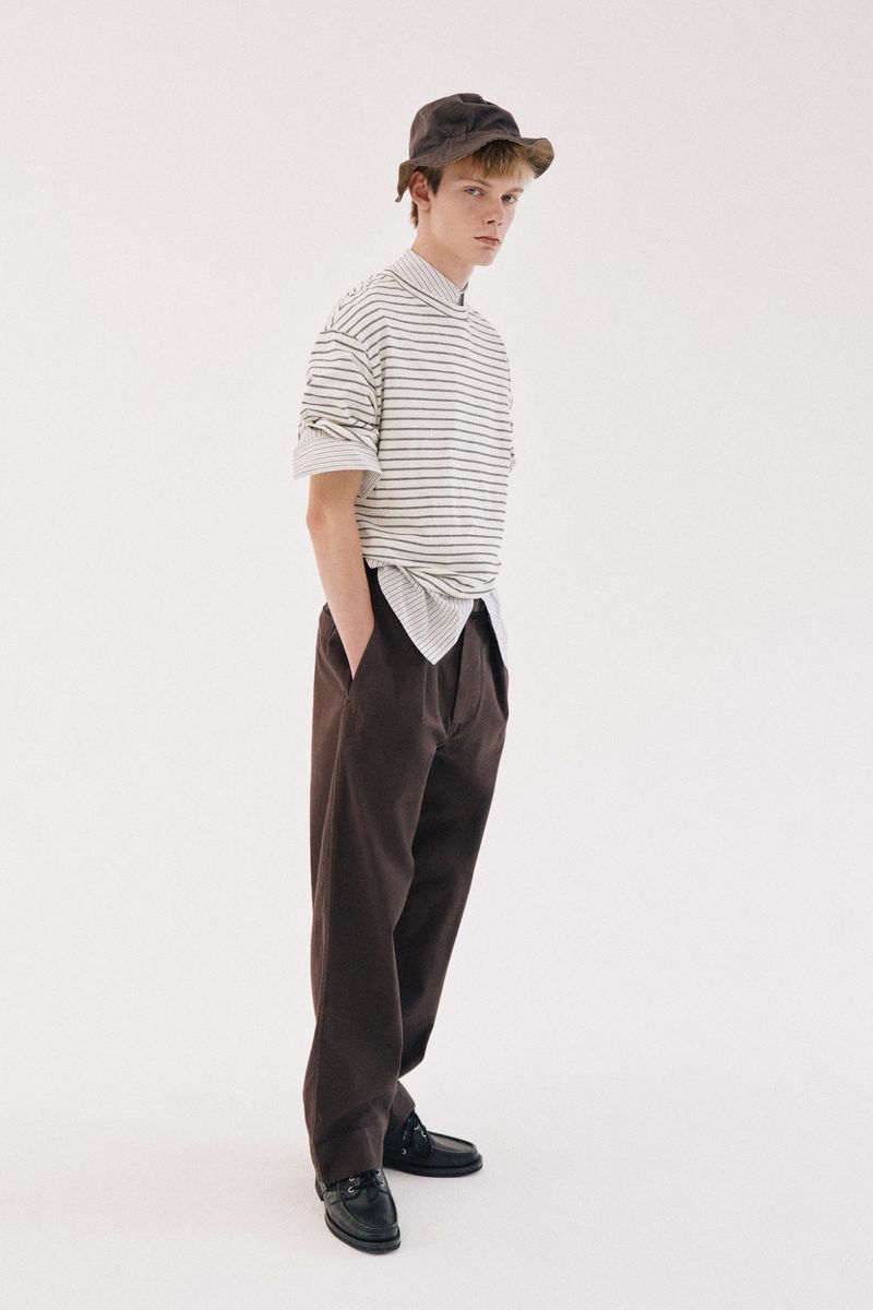 Young male model wearing striped t-shirt and brown trousers with a bucket hat against a white seamless background