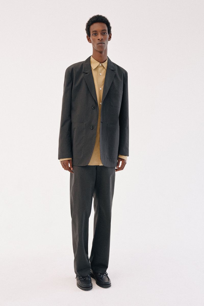 Model standing against a white background wearing a dark suit jacket and trousers over a tan collared shirt