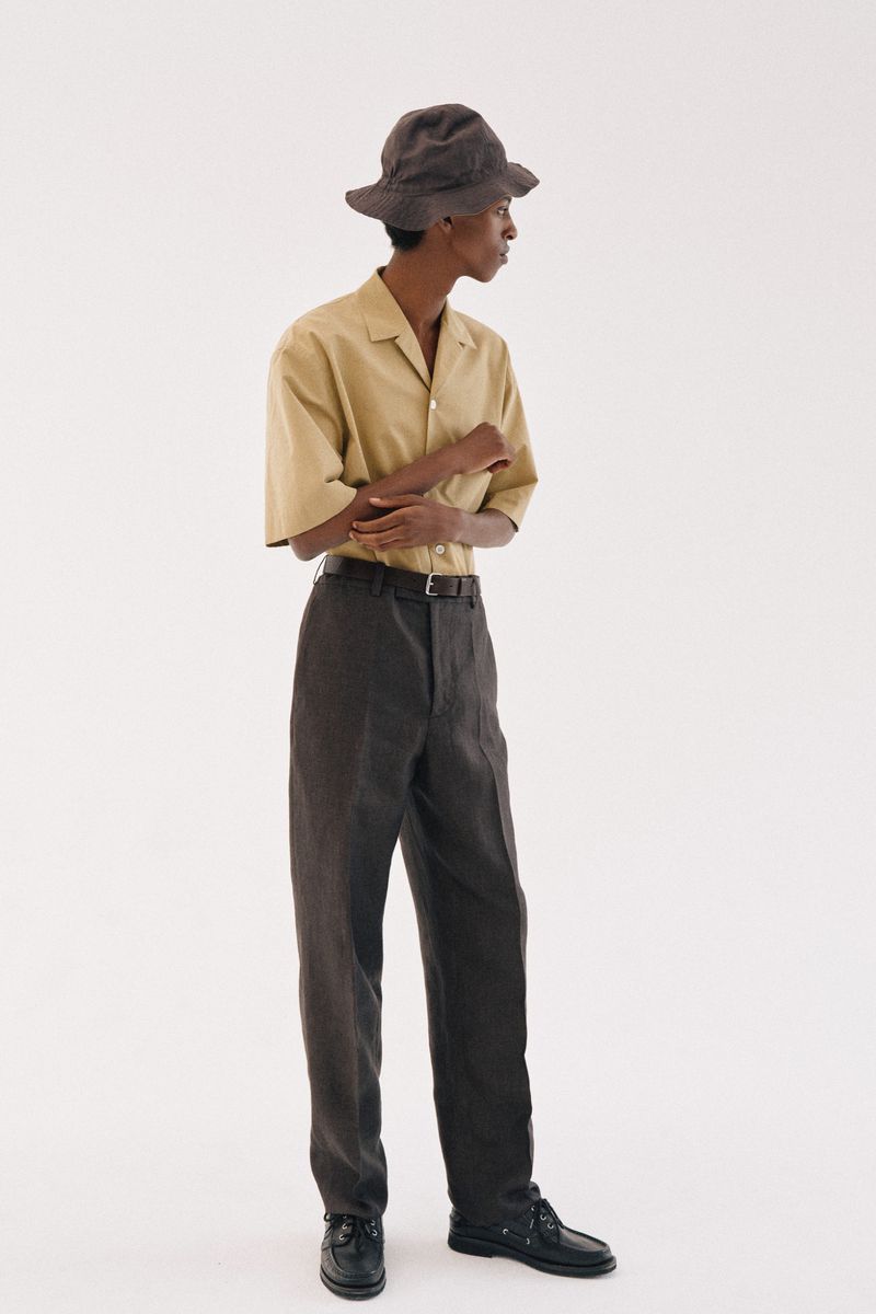 Model wearing a tan short-sleeve shirt, dark trousers, bucket hat, and black shoes against a plain white background.