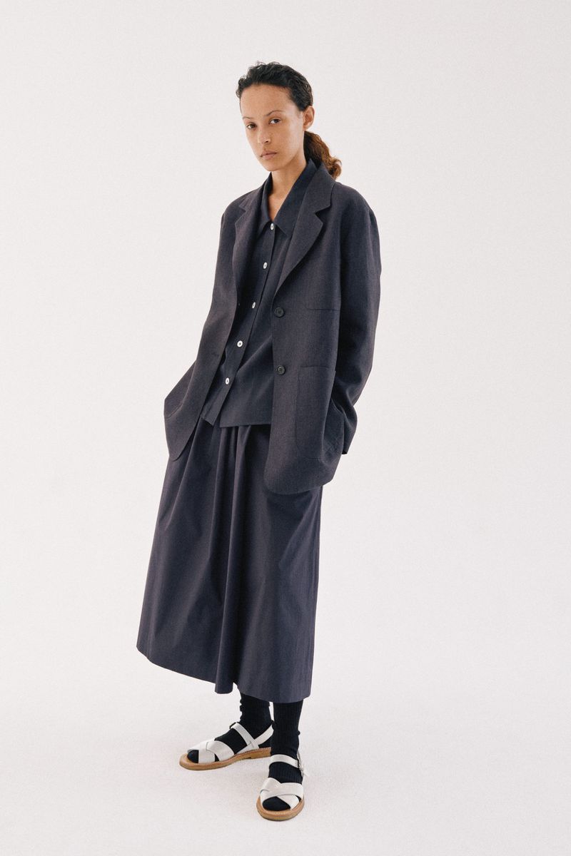 Model standing against a plain white background wearing a dark oversized blazer, matching shirt, and skirt with sandals.