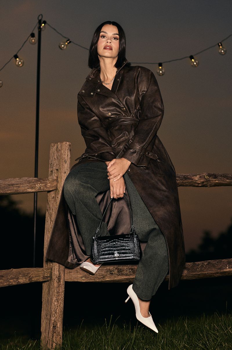 Woman in dark leather coat sitting on a wooden fence holding a black handbag under string lights at dusk