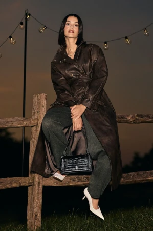 Woman in dark leather coat sitting on a wooden fence holding a black handbag under string lights at dusk