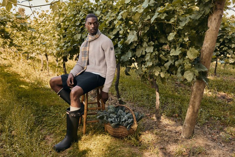 Black man wearing a beige sweater and tall black rain boots seated on a wooden stool inside a vineyard.