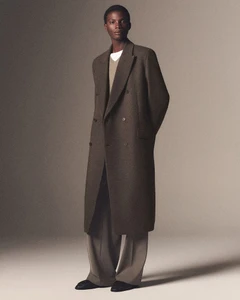 Model wearing a long brown wool overcoat standing against a neutral studio backdrop for COS