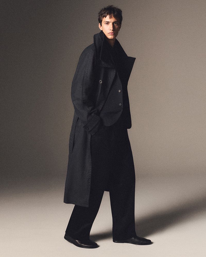 Full-length portrait of a male model wearing a dark long coat and trousers against a neutral grey studio backdrop.