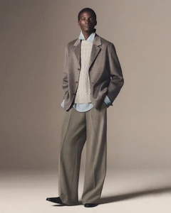 Model standing in neutral-toned outerwear collection by COS wearing a grey jacket over a light blue shirt and trousers.