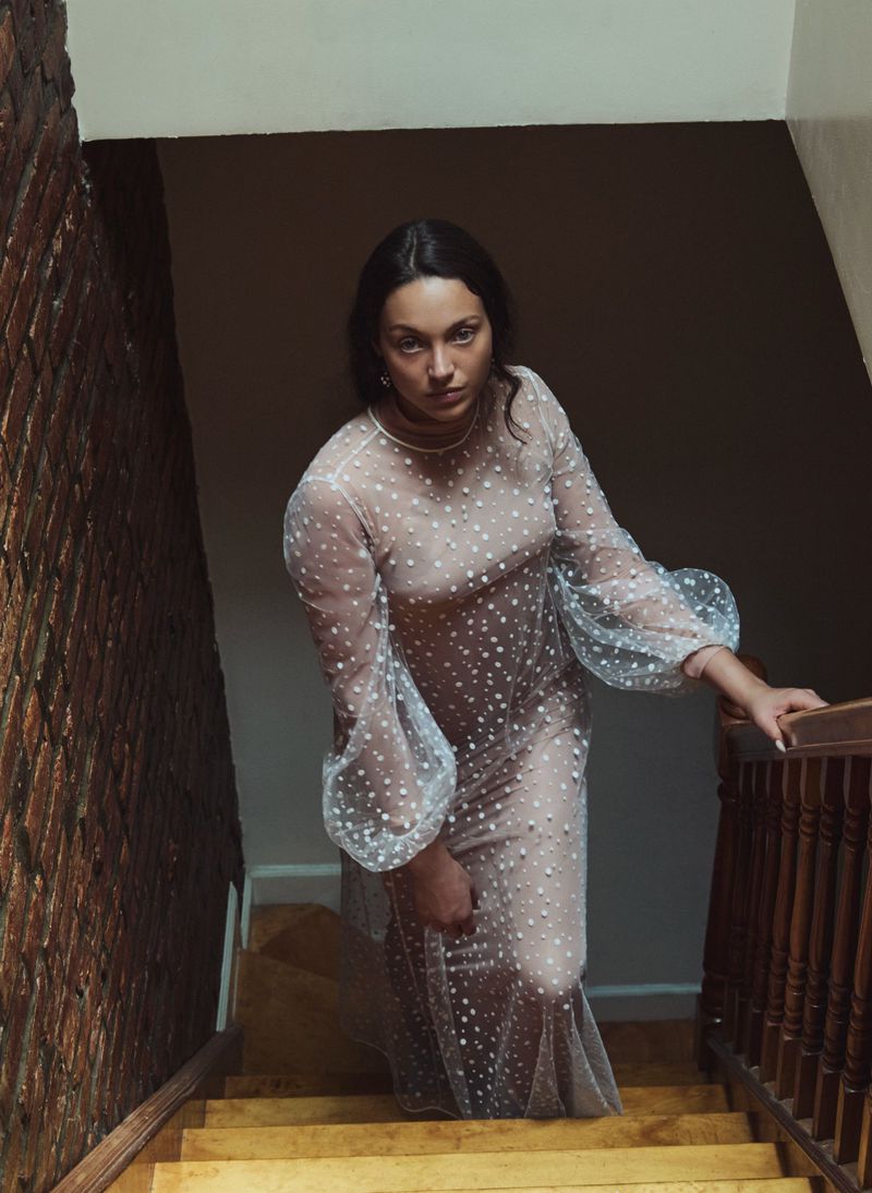 A woman in a sheer, polka-dot gown poses on a staircase with a brick wall, illuminated by soft lighting, creating a sophisticated ambiance.