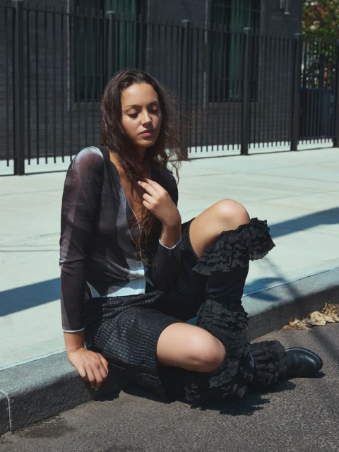 A woman in stylish black clothing sits on a city street curb, with a metal fence behind her in an urban outdoor portrait.