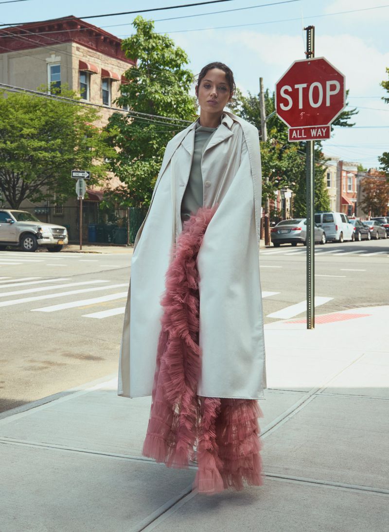 A woman stands on a city sidewalk wearing a beige coat and pink ruffled pants on a sunny day next to a Stop sign on the corner.