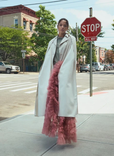 A woman stands on a city sidewalk wearing a beige coat and pink ruffled pants on a sunny day next to a Stop sign on the corner.