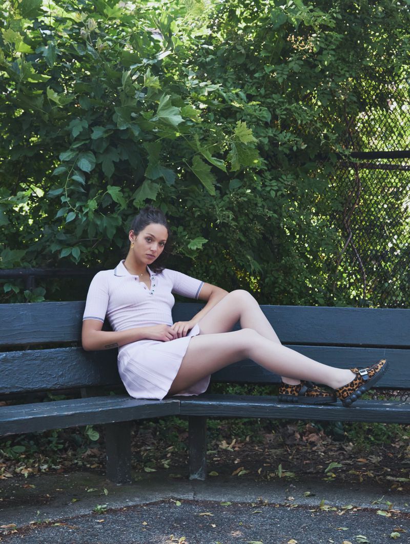 An adult female sits on a park bench with trees in the background. She wears a light-purple tennis outfit and animal-print slip-ons.
