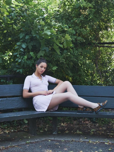 An adult female sits on a park bench with trees in the background. She wears a light-purple tennis outfit and animal-print slip-ons.