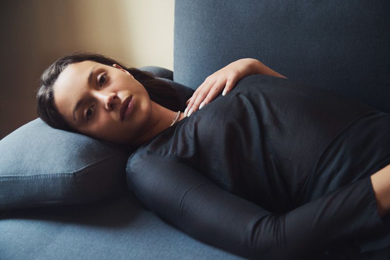 Adult woman in long-sleeved black top resting on blue couch, light skin and dark hair, indoor setting, soft lighting.
