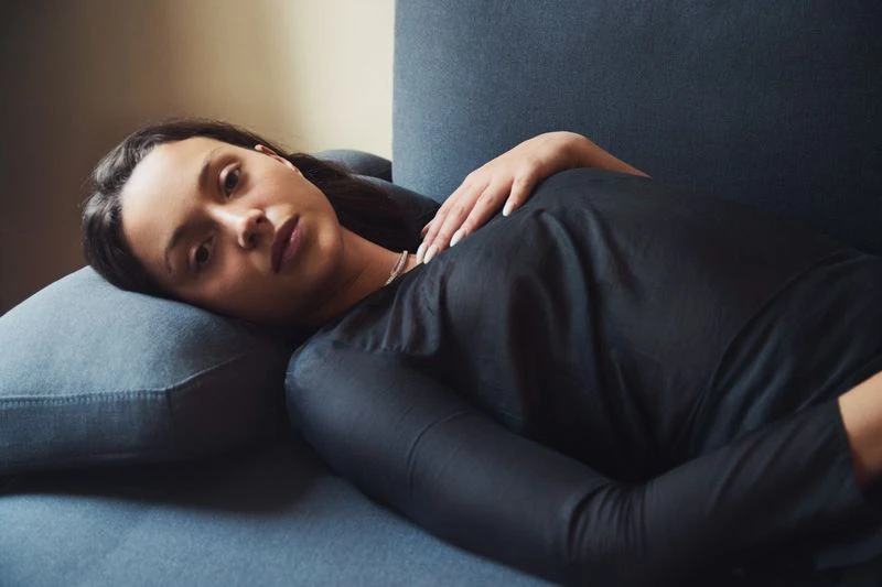 Adult woman in long-sleeved black top resting on blue couch, light skin and dark hair, indoor setting, soft lighting.
