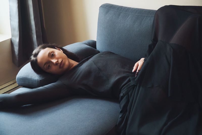 Woman in black dress lies on a blue sofa by a window with curtains, looking at the camera in an indoor setting, soft lighting.