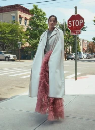Model Jenaye Noah posing in an off-white coat and cascading pink tulle ruffles beside a stop sign on an urban sidewalk.