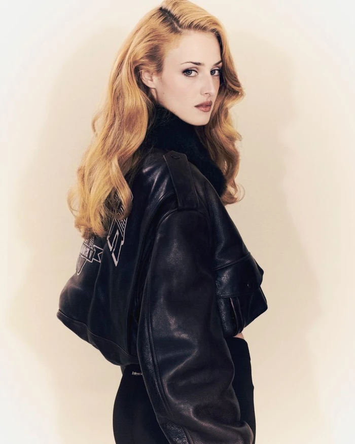 Blonde woman wearing a black leather jacket looking over her shoulder against a plain light background