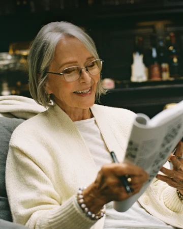 Elderly woman with grey hair wearing glasses and a cream cardigan holding a booklet and pen in an interior setting