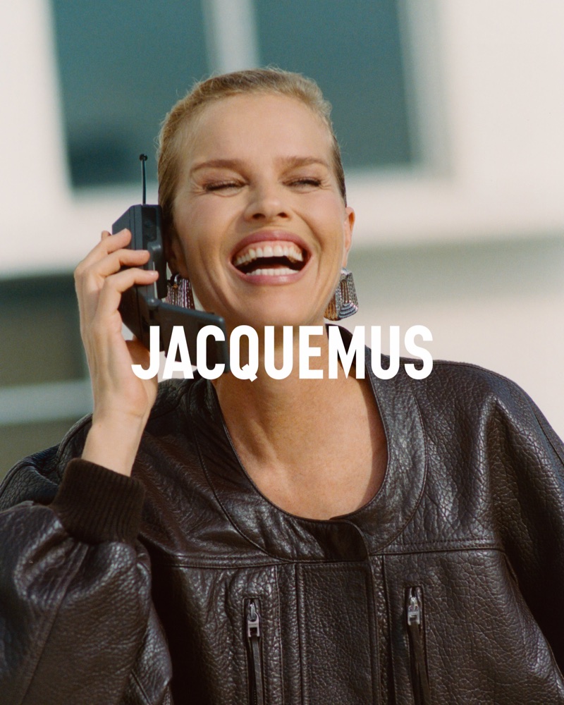 Eva Herzigova laughing while holding a vintage mobile phone, wearing a dark leather jacket by Jacquemus.
