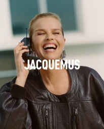 Eva Herzigova laughing while holding a vintage mobile phone, wearing a dark leather jacket by Jacquemus.