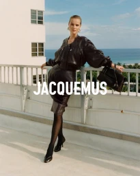 Eva Herzigova leaning on a balcony railing wearing a dark leather jacket and matching skirt against an ocean background